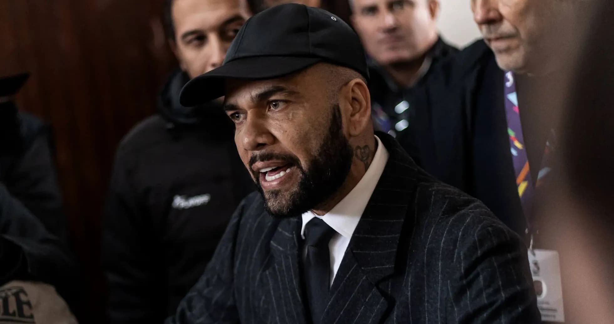 Man in suit speaking with serious expression surrounded by crowd