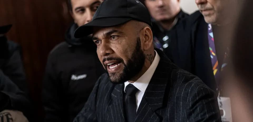 Man in suit speaking with serious expression surrounded by crowd